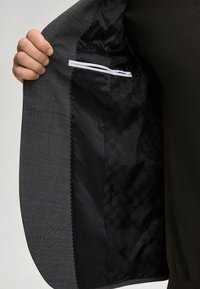 Grauäußeres Stoffblazer mit glattem schwarzen Satinfutter, das ein dezentes Muster aufweist und einen weißen Akzentstreifen in der Innentasche hat.