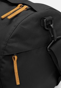 Black fabric duffel bag with textured surface, featuring two leather zipper pulls. Accents include a black clip and adjustable strap detail.