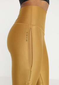 Leggings elasticizzati a vita alta, di colore dorato, con zip laterale e stampa numerica discreta lungo la cucitura.