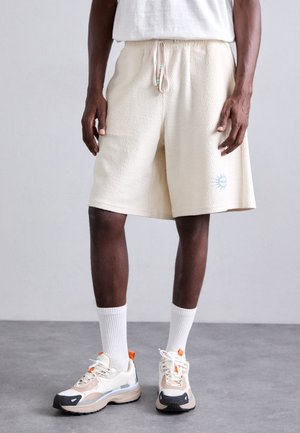 Shorts texturés en gaufre de couleur crème avec une taille élastique et un cordon de serrage. Présente un petit graphique de soleil bleu sur la jambe inférieure.