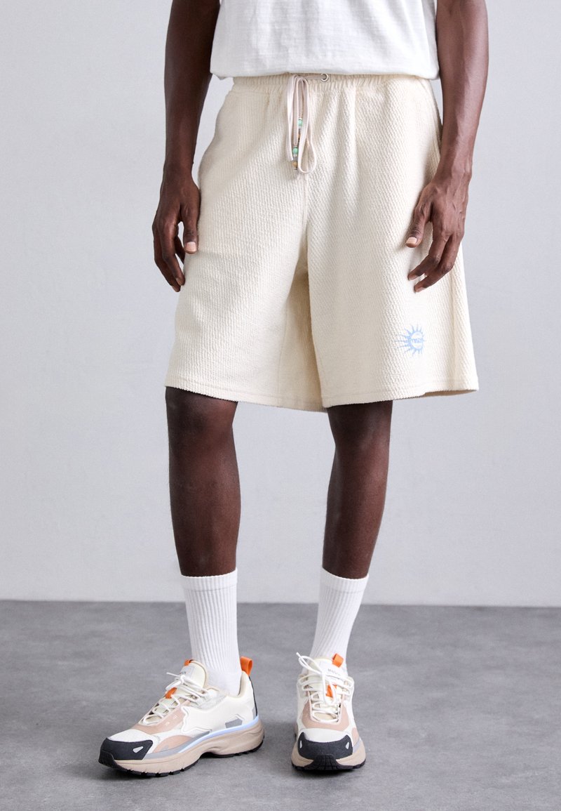 Kremfargede shorts med vaffeltekstur, elastisk midje og snorlås. Har et lite blått solgrafikk på nedre ben.