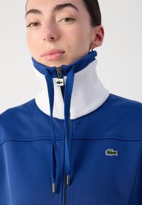 Casaco azul de gola alta com gola plissada branca, fecho com zíper e detalhe de logótipo. Tecido suave, detalhes com cordão e design estruturado.