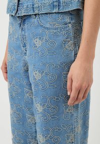 Niebieskie dżinsowe spodnie z beżowymi haftowanymi kwiatowymi i spiralnymi wzorami, noszone z dopasowaną kurtką dżinsową, pokazujące bok z dłonią opartą obok nogi.