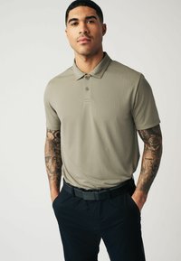 Hellolivgrünes Poloshirt mit kurzen Ärmeln und strukturiertem Stoff, drei Knöpfen am Kragen, kombiniert mit schwarzen Hosen und einem Gürtel.