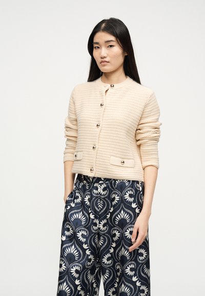 Γυναίκα με μακριά σκούρα μαλλιά που φοράει εκρού υφασμένο πουλόβερ με κουμπιά και σκούρο μπλε παντελόνι με λευκά σχέδια από λουλούδια και φύλλα.