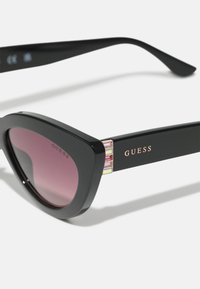 Lunettes de soleil noires en forme de chat avec des verres violets dégradés. Elles présentent une finition brillante et des accents décoratifs sur les branches. Le logo de la marque est visible.