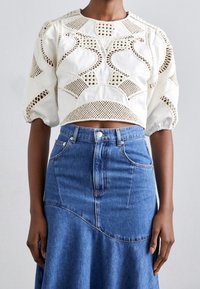 Blouse blanche cropped avec des motifs découpés complexes et des manches bouffantes, associée à une jupe en denim bleue à taille haute avec des coutures visibles.