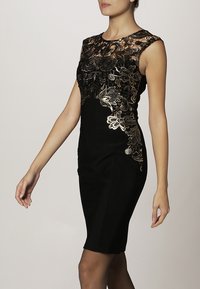 Svart figursydd klänning med en överdel i blommig spets med gyllene accenter. Har rund halsringning och är ärmlös. Slät textur.