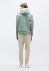 Φαρδιά ανοιχτοπράσινη φούτερ με κουκούλα και ριμπ μανσέτες· συνδυασμένη με μπεζ φόρμα με πλαϊνή τσέπη και λευκά αθλητικά παπούτσια.