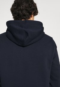 Osoba o ciemnych, kręconych włosach, ubrana w granatową bluzę z kapturem, pokazana od tyłu na jasnym, jednolitym tle.