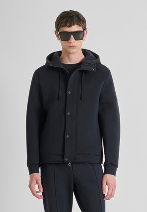 Schwarzes Kapuzensweatshirt mit durchgehendem Knopfverschluss, Kordeln und Seitentaschen. Glatte Textur und modernes Design. Kombiniert mit dunklen Hosen und Sonnenbrille.