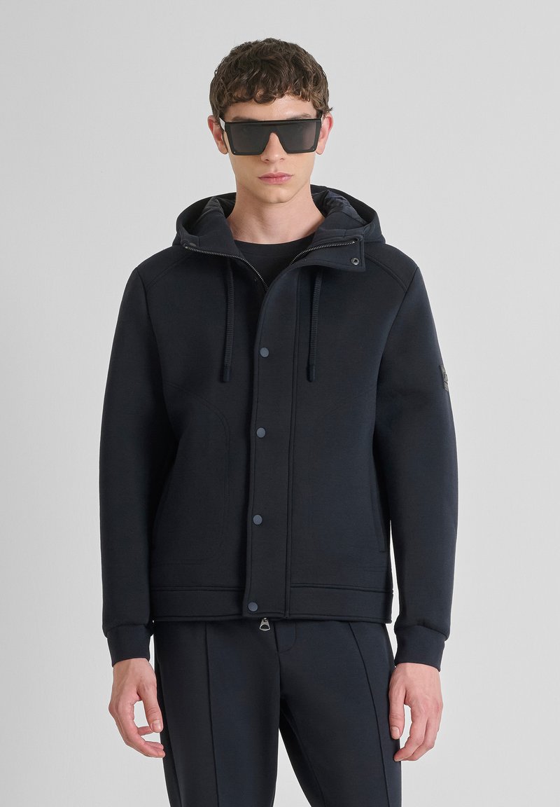 Zwart capuchon sweatshirt met een knoopsluiting aan de voorkant, koordjes en zijzakken. Glad textuur en modern ontwerp. G gedragen met donkere broeken en een zonnebril.