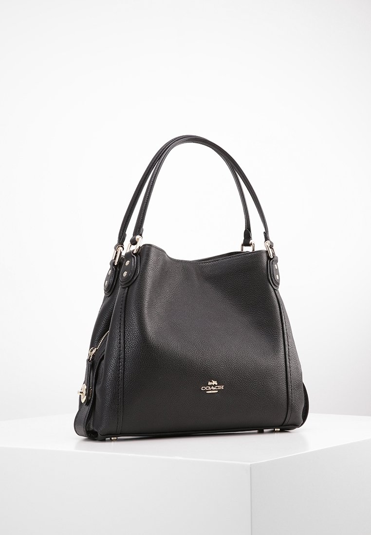 Sac à main Coach en cuir noir avec poignées doubles, poche latérale zippée et logo argenté sur le devant, posé sur une surface blanche.