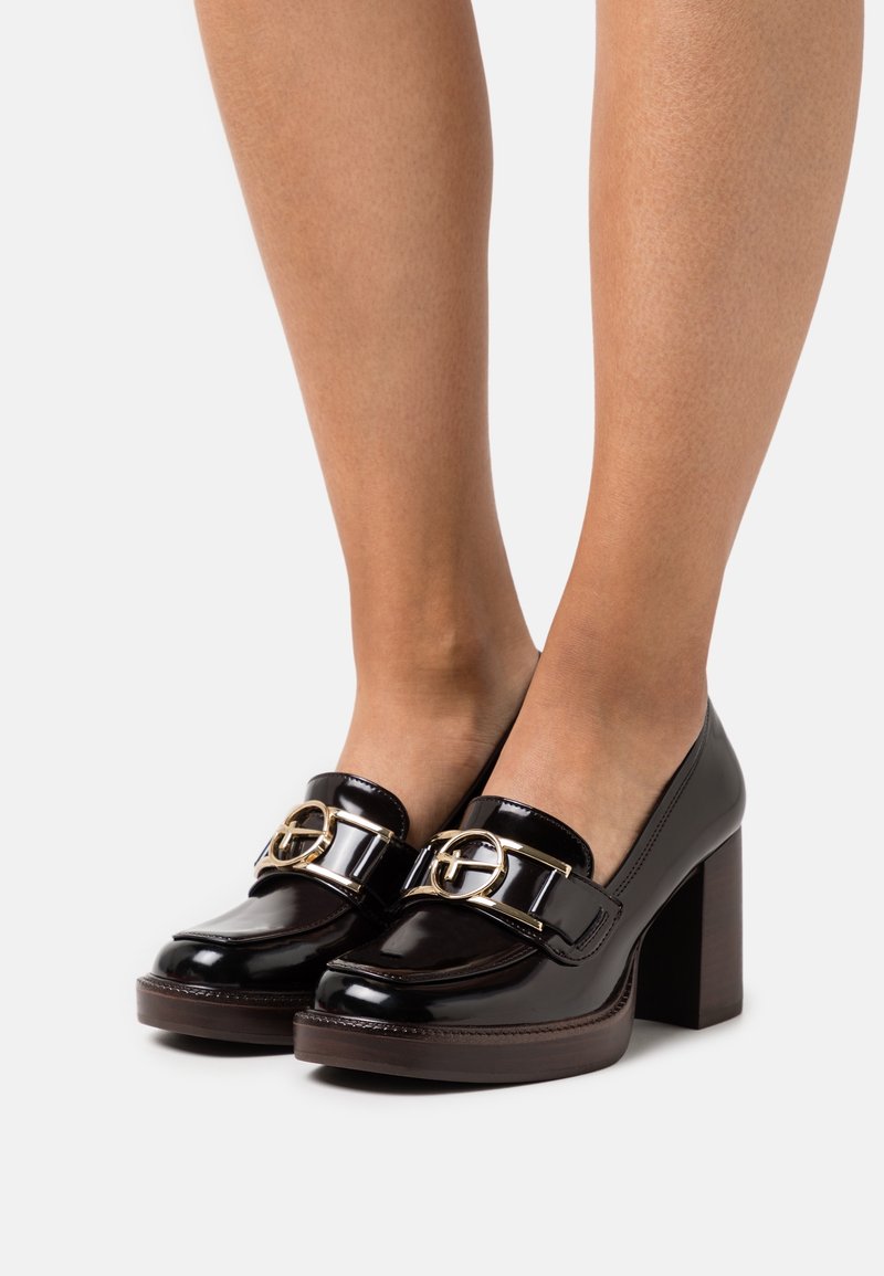 Mocassins en cuir verni noir avec un talon carré et des détails en métal doré. Présentent une texture lisse et un design à bout arrondi.
