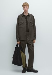 Camicia marrone con bottoni e due tasche frontali, pantaloni abbinati e scarpe nere. Tiene una borsa a tracolla scura rivestita di tessuto verde.