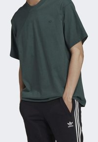 Person, der ein dunkelgrünes Kurzarmshirt und schwarze Adidas-Trainingshosen mit weißen Streifen trägt, die Hände in den Taschen, vor einem schlichten Hintergrund.