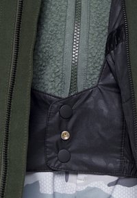 Strato esterno verde con zip, foderato in fleece, tessuto interno nero, chiusura con bottoni e camo grigio a motivo sull'orlo.