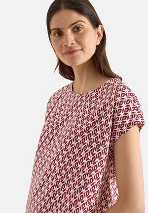 Kvinde med langt brunt hår iført en kortærmet top med et rødt og hvidt diamantmønster, der kigger let til siden med et neutralt udtryk.