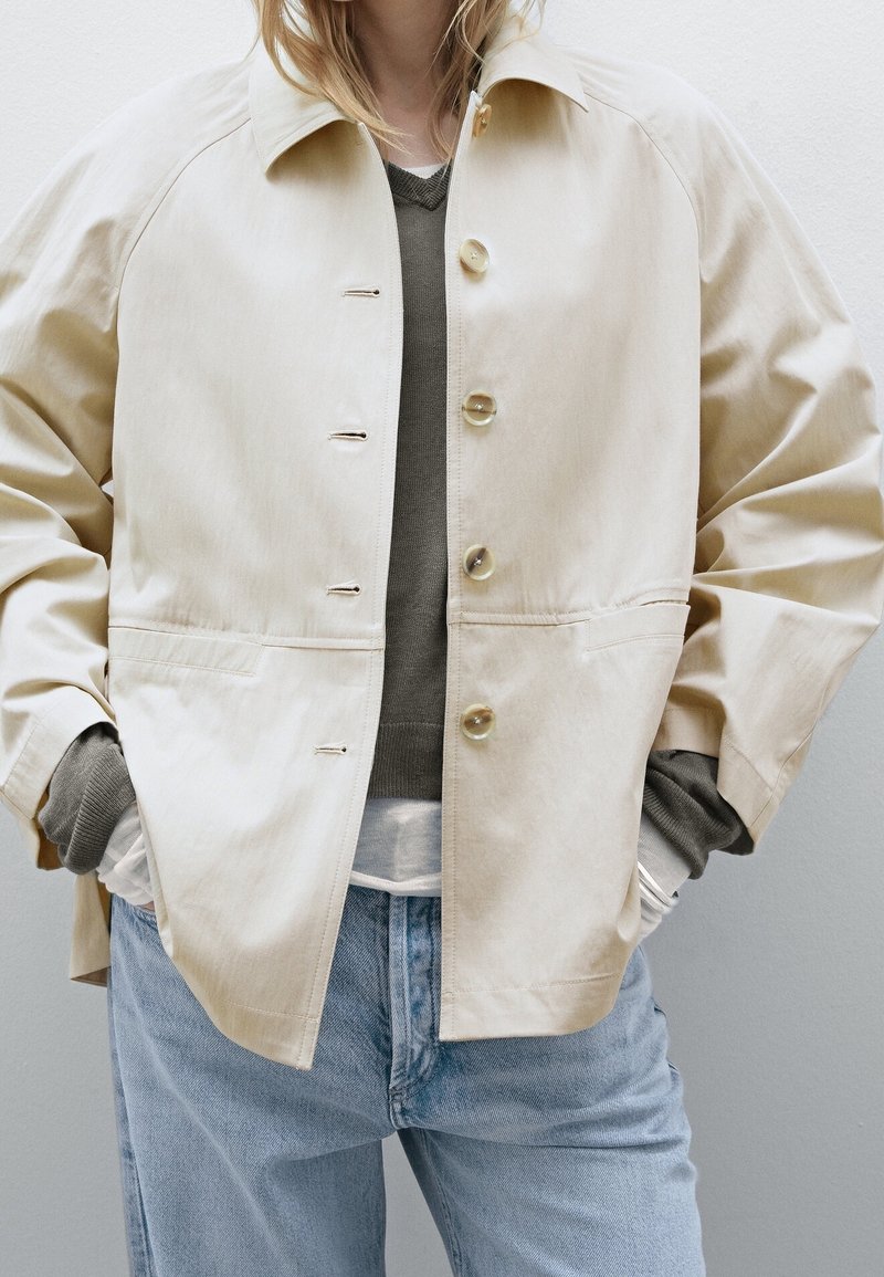 Persoon in een beige knoopjasje over een grijze trui en een wit shirt, met lichtblauwe jeans en handen in de zakken van het jasje.
