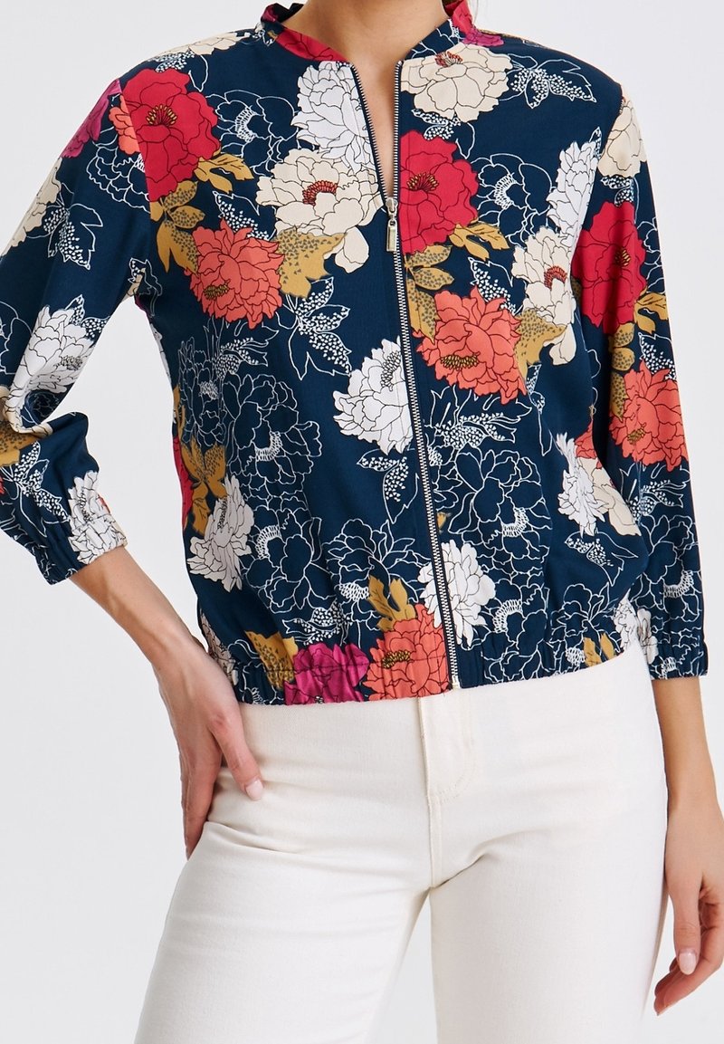 Chaqueta con cremallera y estampado floral que presenta un fondo azul marino, patrones de flores multicolores y elásticos en el dobladillo y las mangas. Tela ligera.