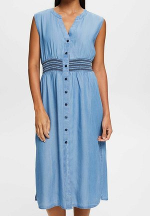 Ärmelloses Denim-Midikleid mit geraffter Taillendetail, Frontknopfleiste und leichtem V-Ausschnitt, getragen von einer stehenden Person.