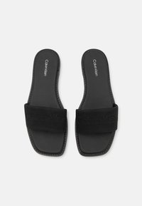Black slide sandals featuring a wide fabric strap with Calvin Klein branding, smooth synthetic sole, and square toe design.