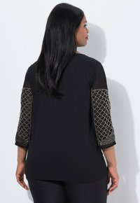 Zwarte blouse met driekwartmouwen, gemaakt van een transparante stof en gouden geschulpte borduursels. Zachte textuur; losse pasvorm.