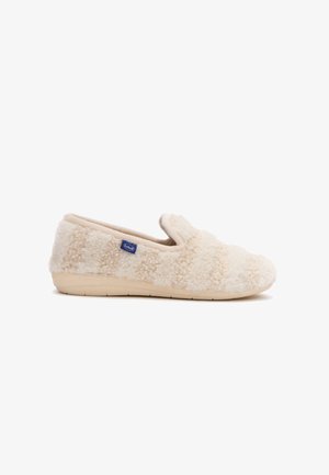 Zapatilla de felpa color crema con parte superior texturizada, tacón bajo, suela suave y una pequeña etiqueta azul de Scholl en el lateral.