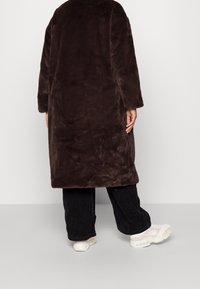 Bruine faux fur jas met een relaxte pasvorm, lange mouwen en een gladde textuur. Draagt over zwarte corduroy broek en witte sneakers.