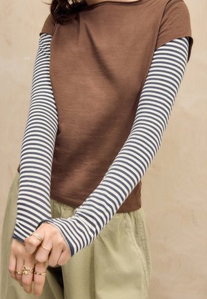 Personne portant un haut marron à manches courtes superposé sur une chemise à manches longues rayée bleu marine et blanc avec un pantalon kaki, bras croisés vers l’avant.