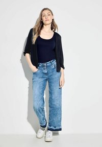 Svart öppen cardigan, marinblå tanktop, ljusblå vida jeans med fransad fåll, tillsammans med vita sneakers, mot en neutral bakgrund.