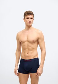 Άνδρας σε φόρμα με κοντά σγουρά μαλλιά, φοράει μπλε ναυτικό εσώρουχο Lacoste με κόκκινο λογότυπο στη μέση, με φόντο ανοιχτό.