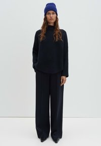 Schwarzer gerippter Pullover mit hohem Kragen, kombiniert mit weit geschnittenen schwarzen Hosen und einer strukturierten blauen Strickmütze. Glattes, minimalistisches Design.