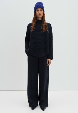 Zwart geribbelde trui met een hoge hals, gecombineerd met zwarte wijde broek en een blauw gebreide muts met textuur. Glad, minimalistisch ontwerp.