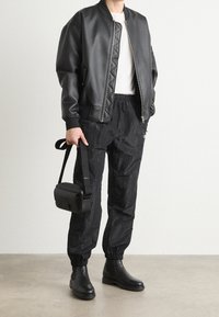 Černá bomber bunda s prošívanou strukturou, bílý tričko pod ní, černé lehké kalhoty, černé kotníkové boty a malá černá kabelka přes rameno.