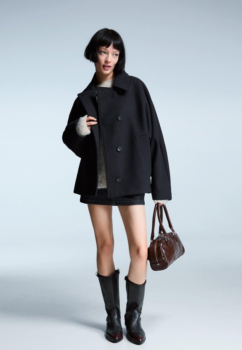 Črn oversized plašč z velikimi gumbi, oblečen preko kremastega puloverja. V kombinaciji z jeans krilom, črnimi škornji do kolen in rjavim ročnim torbico.