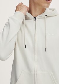 Hvit zip-up hettegenser med myk tekstur, justerbar hette, svarte snorer og en liten 'blend'-logo på brystet. To frontlommer.