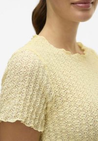 Gul laces topp med korte ermer, scalloped halslinje og et teksturert mønster med åpne vever og subtile detaljer.