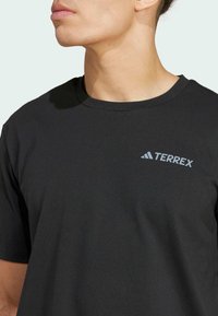 Black cotton t-shirt featuring a round neck and short sleeves, with a grey "TERREX" logo on the left chest. Smooth texture, casual design.
