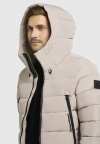 Bežna pufasta jakna z velikim kapuco, zadrgo spredaj in dvema stranskima žepoma. Ima quiltasto teksturo in črne kovinske dodatke.