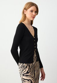 Zwarte geribde cardigan met een V-hals, voorzien van gouden knopen aan de voorkant. Draag het over een beige rok met zwarte zebraprints.