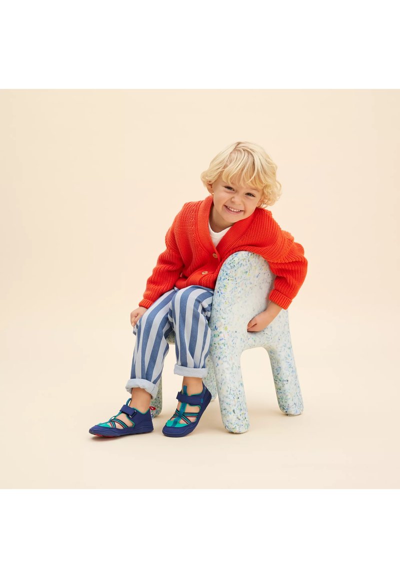 Bambino che indossa un cardigan rosso a maglia, pantaloni a righe blu e scarpe blu con dettagli verdi, seduto su una sedia maculata contro uno sfondo neutro.