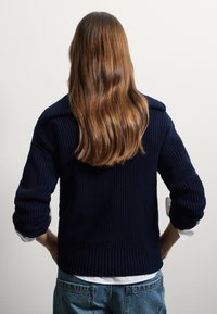 Pull en maille navy avec texture côtelée, coupe oversize et large col. Porté par-dessus une chemise blanche, associé à un jean en denim bleu clair.