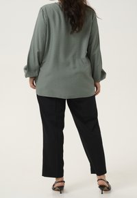Blouse à manches longues, verte, avec des pois texturés, associée à un pantalon noir ample et des sandales à talons noires.