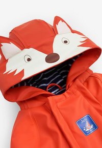 Veste imperméable orange vif avec une capuche en forme de tête de renard, comprenant des oreilles et des yeux. Intérieur doublé de tissu rayé bleu marine et blanc.