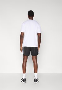 Camiseta blanca de manga corta, pantalones cortos deportivos negros con rayas blancas a los lados, zapatos deportivos negros, vistos desde atrás sobre un fondo liso.