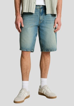 Mann trägt hellblaue Jeansshorts, weiße Socken und beigefarbene Turnschuhe mit weißen Streifen, steht vor einem schlichten Hintergrund.