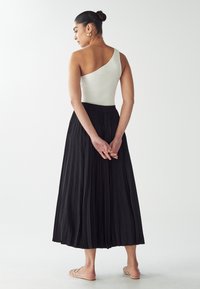 Black pleated maxi skirt with an elastic waistband, paired with a ribbed one-shoulder white top. Minimal accessories and flat sandals.