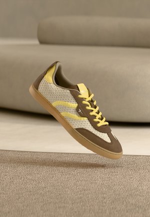 Zapatilla marrón y crema con detalles amarillos, parte superior de tela de malla, suela de goma y rayas contrastantes en los lados, mostrando un diseño moderno.