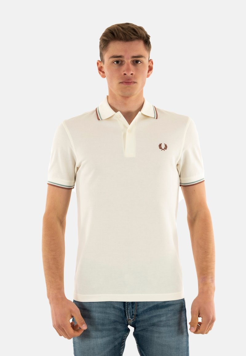 Hvid polo-shirt med en ribbet krave med grønne og røde striber. Skjorten har et lille broderet emblem på venstre bryst.